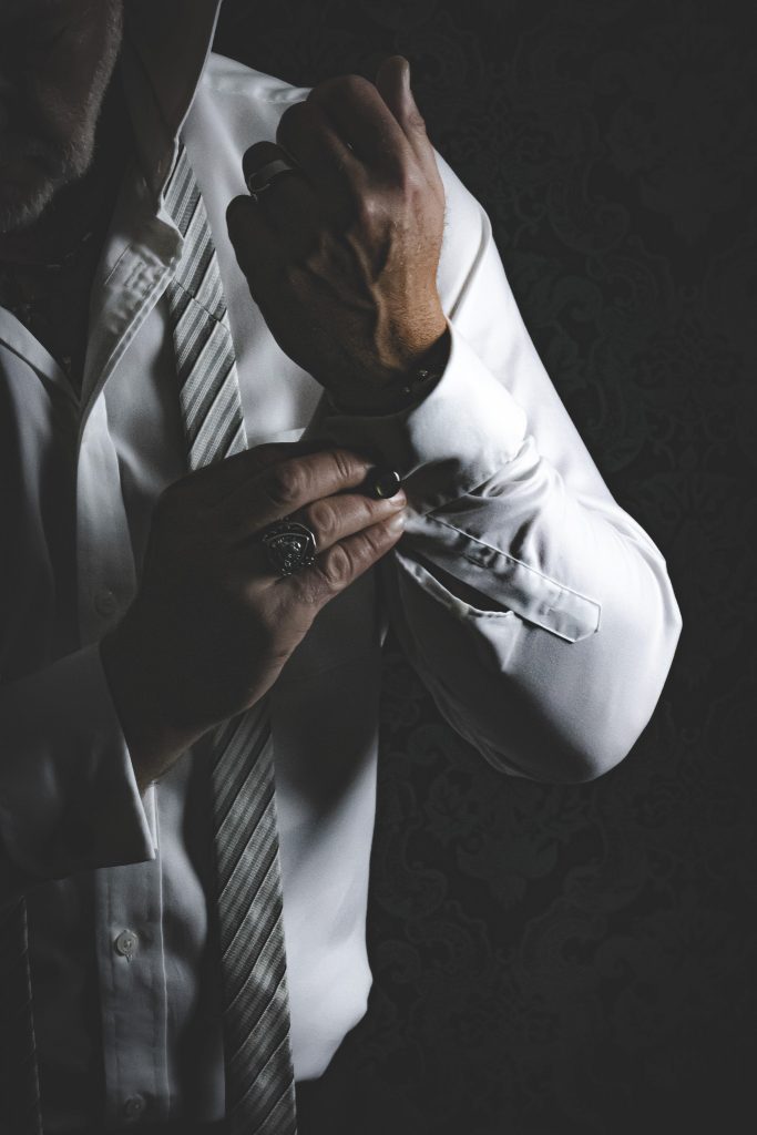 Ein männliches Model im halbdunkel. Der Bildschnitt zeigt sein gedankenverlorens anlegen der Manschettenknöpfe. Sein Auftritt wird von seiner Elegeganz und seinem starkem Selbstbewusstsein geprägt.